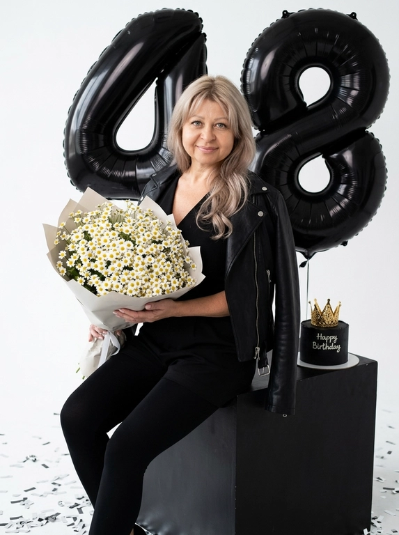 Максимально реалистичная фотография в стиле luxury birthday editorial, элегантная и торжественная студийная съемка премиального уровня.
Полный рост. женщина сидит на матовом черном кубе, уверенно и расслабленно удерживая перед собой большой роскошный монобукет цветов в упаковочной бумаге. Взгляд направлен прямо в камеру, легкая полуулыбка создает спокойное и уверенное настроение кадра.
Макияж вечерний и элегантный: с мягкой растушевкой, густые ресницы.
Одежда: черный облегающий ромпер с короткими шортами, подчеркивающий силуэт. Поверх небрежно накинута черная кожаная куртка-косуха на одно плечо. Мягкая фактура кожи с естественными бликами и складками материала.
Локация: минималистичная белая фотостудия. На заднем плане расположены большие черные фольгированные шары в форме цифр. На полу рассыпано серебристое конфетти, рядом с кубом стоит небольшой черный праздничный торт с золотой надписью «Happy Birthday» и декоративной короной.
Освещение: профессиональный студийный свет с мягким рисующим источником спереди и легким контровым светом, создающим объем и мягкие естественные тени.
Съемка на полнокадровую камеру, объектив 50mm. Модель, монобукет и торт находятся в идеальном фокусе, задний план слегка размыт.
Гиперреалистичная текстура кожи без пластиковой ретуши: видны поры, естественный микрорельеф кожи, текстура рук и ног, натуральные блики на коже и волосах.
Высокая детализация лепестков цветов, упаковочной бумаги букета, текстуры кожи куртки, отражений на фольгированных шарах и фактуры торта.
RAW photo, ultra photorealistic, luxury birthday editorial, studio photography, natural skin texture, realistic lighting, subtle film grain, high detail, 8K, vertical 2:3.
Без AI-артефактов, пластикового эффекта кожи, пересветов, HDR, неестественных цветов, деформации рук, ног и пальцев.

Шары в форме цифр: 48

Какие цветы в букете?: Ромашки 

Доп. характеристики: Ноги в лосины