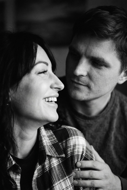 Сгенерируй Черно-белое фото, кинематографичная фотография пары, снятая крупным планом. девушка находится слева в кадре на переднем плане, повернута в профиль и смеется, не глядя на парня , парень справа на заднем фоне смотрит на глаза девушки задумчиво и влюбленно, с мягким, нежным, влюбленным выражением лица. свет падает сбоку, создавая мягкие тени на лицах и подчёркивая черты лица . фон размытый , темный , без четких деталей , создававший атмосферу уюта фотография выглядит как кадр из романтического фильма или личный момент пойманный камерой формат фото