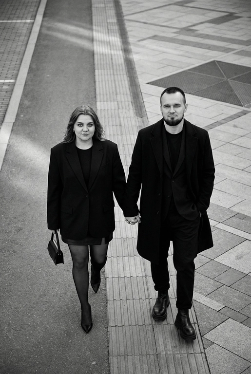 Черно-белая художественная фотография, ультрареализм, 8K, профессиональная съёмка; (оба): в кадре мужчина и женщина; каждый персонаж жёстко привязан к своему загруженному референсу, лица используются напрямую, без генерации, усреднения, подмены или смешения. Вид сверху под углом. Они идут по городской улице, держась за руки; шаг в движении, эффект «пойманного кадра». Смотрим вверх в камеру, выражение спокойное, уверенное, отстранённое.
Полностью чёрные образы modern classic: женщина — оверсайз-пиджак, короткое платье/юбка, плотные колготки, остроносые каблуки, маленькая сумка; мужчина — длинное пальто, свободный костюм, массивные ботинки.
Фон: асфальт, плитка, тактильная дорожка, геометрия.
Контрастный свет, мягкие тени, высокая детализация тканей.
Стиль: black & white, cinematic, high fashion, editorial, minimalism, street photography.
Качество: ultra high resolution, sharp focus, film grain, без ретуши.