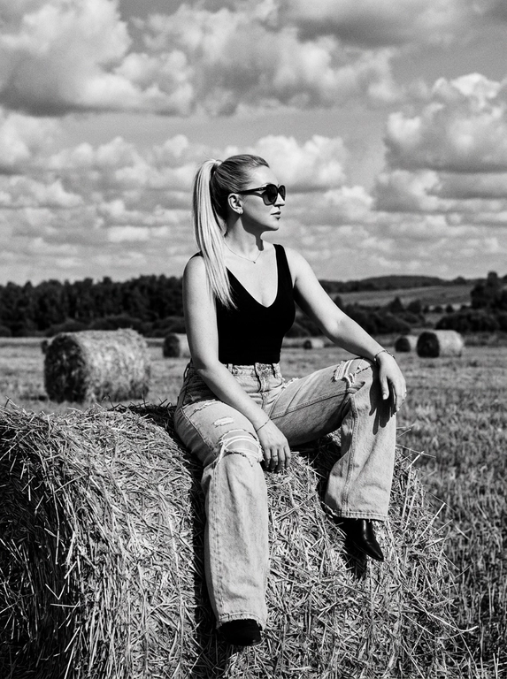 Чёрно-белая high-fashion editorial фотография в сельской местности, женщина с внешностью как на референсе, черты лица не изменять, композиция — более общий план (wide shot), камера на среднем расстоянии, фигура полностью в кадре с окружающим пространством, она сидит на большом тюке сена в поле, корпус слегка развернут, одна нога свободно опущена вниз, вторая согнута, создавая расслабленную, но выразительную позу, руки естественно лежат на колене и на поверхности тюка, взгляд направлен в сторону, профиль, выражение спокойное и уверенное, одежда: чёрный топ с V-образным вырезом и объёмные светлые широкие джинсы с потёртостями и рваными элементами, аксессуары: крупные солнцезащитные очки, минималистичные украшения, фон: открытое поле с несколькими тюками сена на разной глубине, создающими перспективу, линия горизонта ниже центра кадра, чтобы подчеркнуть небо, освещение: жёсткий естественный дневной свет с мягкими облаками, формирует выразительные тени и контраст, стиль: кинематографический, плёночное зерно, высокая детализация, реалистичная текстура кожи и ткани, редакционный fashion стиль, акцент на атмосфере и пространстве кадра


Какая будет прическа?: Гладкие волосы собраны в высокий хвост