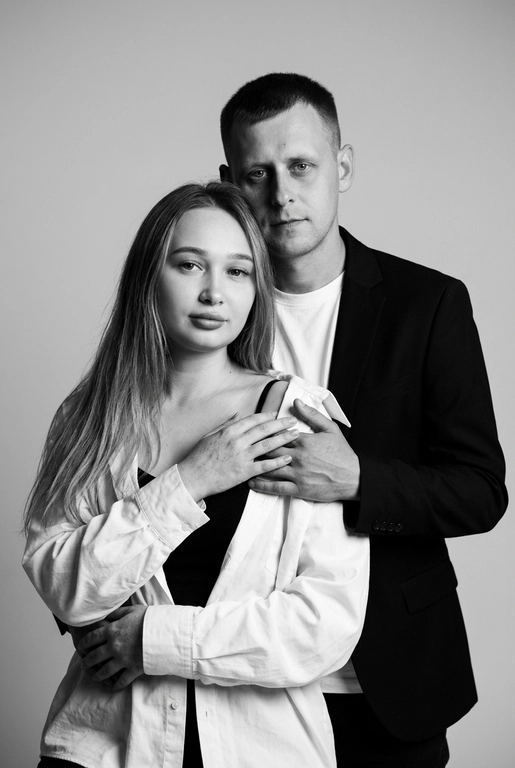 Черно-белый портрет мужчины и женщины в стиле фэшн фотосессии в фотостудии. Не изменяйте лица. Фон нейтральный, однотонный, светло-серый. Атмосфера интимная и спокойная. На переднем плане женщина. Вся композиция выполнена в приглушенной монохромной гамме со студийным освещением, которое подчеркивает текстуру кожи и одежды. Её руки лежат на груди мужчины. Мужчина обнимает женщину сзади, положив свою руку поверх её руки.

Она одета в черный топ, белую оверсайз рубашку. Мужчина одет в  классическом черном пиджаке и белой футболке. Она более худая , вес 50 килограммов,  у него менее торчат уши