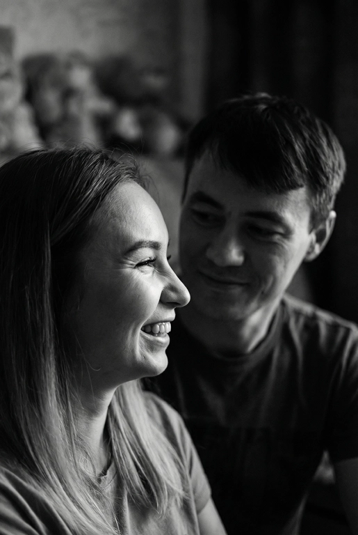   Сгенерируй Черно-белое фото, кинематографичная фотография пары, снятая крупным планом. девушка находится слева в кадре на переднем плане, повернута в профиль и смеется, не глядя на парня , парень справа на заднем фоне смотрит на глаза девушки задумчиво и влюбленно, с мягким, нежным, влюбленным выражением лица. свет падает сбоку, создавая мягкие тени на лицах и подчёркивая черты лица . фон размытый , темный , без четких деталей , создававший атмосферу уюта фотография выглядит как кадр из романтического фильма или личный момент пойманный камерой формат фото