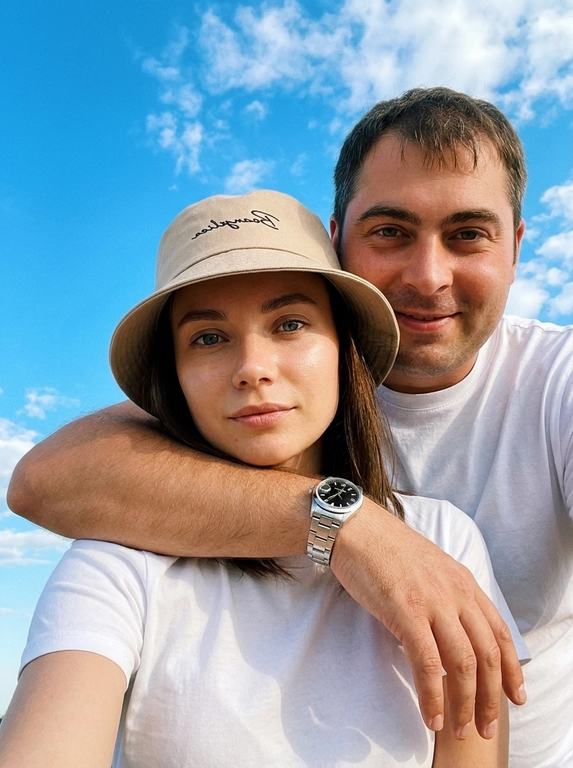 Кинематографичный кадр крупным планом (selfie style), пара в объятиях (классическое "обнимашки сзади") на фоне ярко-голубого неба с облаками.  Женщина на ней бежевая панама с брендовой надписью. Её кожа выглядит сияющей, подсвеченной мягким естественным солнечным светом. Выражение лица спокойное, глубокий взгляд, направленный прямо в камеру. Мужчина стоит строго позади девушки и обнимает её сзади, одна его рука обнимает её за плечи/шею спереди — предплечье лежит поперёк её ключиц, кисть свободно свисает с её плеча, на запястье классические серебряные часы с черным циферблатом. Мужчина в белой футболке, выражение лица доброжелательное, его взгляд прямо в объектив. Ракурс сверху, камера сфокусирована на лице девушки. Высокий контраст, насыщенные цвета, мягкая прорисовка деталей кожи. Эффект пленочной фотографии, легкое зерно, атмосфера летнего дня, эстетика социальных сетей. Телефона не видно в кадре.