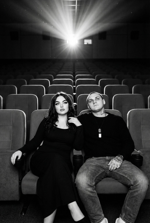  Кинематографичный чёрно-белый портрет пары в пустом кинозале.
Женщина и мужчина сидят рядом в мягких креслах кинотеатра.
кадр с глубоким пространством и пустыми рядами кресел позади них. Женщина черном длинном обтягивающем платье, поза женственная и спокойная, одна рука на подлокотнике.
Мужчина в черном свитере и в джинсах, сидит расслабленно.
Над ними яркий луч проектора, и выразительные блики.
