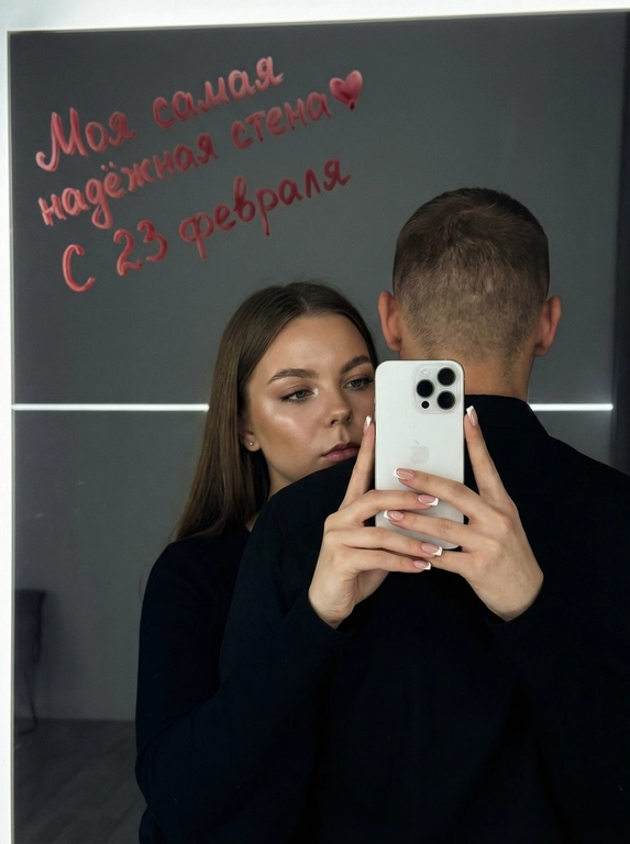 Ультрареалистичное кинематографичное зеркальное селфи пары. Строго сохранить лица из референса без изменений: возраст, пропорции, форму глаз, носа и губ, текстуру кожи, линию подбородка. Не изменять черты лица.

Локация: Серый лофт, low-key атмосфера.

Композиция: мужчина стоит спиной к камере, его лицо полностью не видно (back fully facing camera); за его спиной женщина обнимает его и снимает зеркальное селфи. Она держит белый смартфон Apple iPhone 16 Pro двумя руками. Фокус на руках и телефоне, френч квадратный маникюр. Физически корректное зеркальное отражение, без искажений..

Образ девушки: безупречный макияж, лёгкое сияние кожи.

Свет и стиль: подсветка от зеркала сверху-спереди, мягкие тени, тонкая горизонтальная световая линия на фоне. 50-85 мм, f/2-2.8, малая ГРИП; киноконтраст, плёночное зерно, high-end editorial, 8K.

Сцена полностью показана через отражение в зеркале, единственный источник изображения  — это отражение.

Ваша надпись красной помадой на зеркале: Моя самая надёжная стена❤️ С 23 февраля

Ваша одежда: Мужчина и женщина одеты в чёрной рубашке;