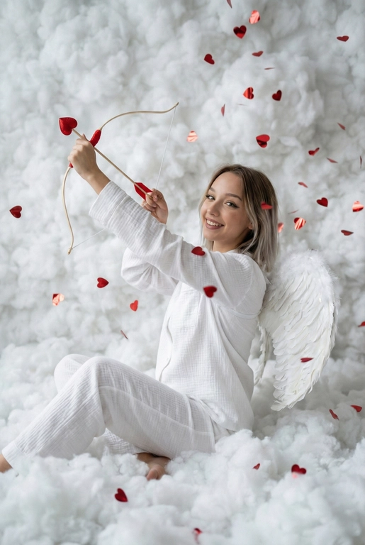 Использовать реальные черты лица человека с референса, естественную текстуру кожи и настоящий цвет и длину волос. Личность должна оставаться неизменной.

Y2K. Сказочная сцена с купидоном, сфотографированная как реальный момент.
Человек сидит среди плотных, пушистых белых облаков, которые полностью заполняют передний, средний и задний план, образуя бесшовную белую среду с многослойной глубиной.

Поза вполоборота: корпус слегка повернут вбок, одно плечо выдвинуто вперед, как будто человек натягивает и отпускает небольшой лук с красной стрелой в форме сердца; руки подняты с легким напряжением, указывающим на движение при выстреле.

Несмотря на поворот тела, голова повернута к объективу, взгляд направлен прямо в камеру. Выражение лица радостное и игривое, с мягкой улыбкой.

Наряд — простая белая пижама: свободная рубашка и брюки из муслина.
На спине — крылья из белых перьев, настоящие и фактурные, с четкими бликами по краям каждого пера.

Вокруг объекта в воздухе хаотично летают только конфетти в форме сердечек — маленькие красные сердечки из глянцевой фольги, расположенные на разном расстоянии: часть в переднем плане, часть растворяется среди облаков.

Без рассеянного света, без мягкого свечения, без студийного глоу.
Текстура кожи реалистичная и детализированная, без сглаживания и без фильтров.
Тонкая зернистость, волшебное, но искреннее ощущение Y2K-редакции — выглядит как фотография, а не визуализация.
