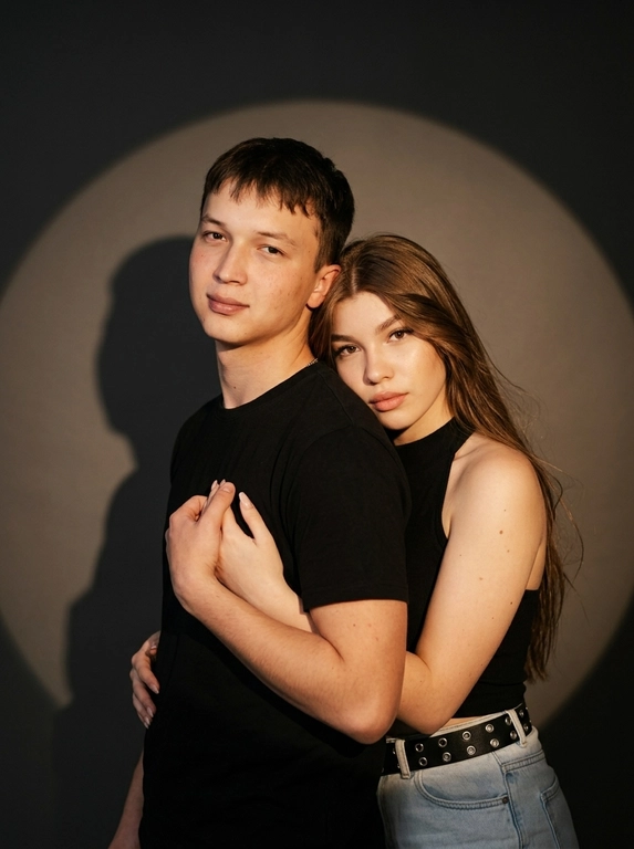 Фотореалистичный студийный портрет пары в драматичной Y2K fashion-эстетике. Мужчина стоит на переднем плане, ближе к камере, снят по грудь, крупнее в кадре. Его корпус слегка повернут в пол-оборота, одно плечо ближе к объективу, поза спокойная и уверенная. Женщина стоит позади него, немного ниже по уровню за счёт наклона корпуса, частично скрыта его плечом. Она мягко обнимает его за шею и плечо, её руки лежат спереди у его груди и ключиц. Мужчина одной рукой аккуратно держит её кисть. Её лицо видно из-за его плеча, близко к нему, поза выглядит интимно, дерзко и editorial, без чрезмерной романтичности. Женщина выглядит выразительно и гламурно: cat-eye стрелки, губы с чётким контуром, хайлайтер на коже, чёрный корсет и оверсайз-пиджак с открытым плечом. Мужчина одет в чёрный лонгслив. Фон студийный, тёмно-серый, почти чёрный. За парой на стену направлен мощный прожектор, создающий большой круг света позади их силуэтов. Это именно свет от прожектора, падающий на фон, а не нарисованное пятно: круг света мягко размывается по краям и постепенно уходит в темноту. Персонажи частично перекрывают свет от прожектора, поэтому на фоне видны реалистичные жёсткие тени от их тел, рук и плеч. Края кадра затемнены, вокруг светового круга глубокая тёмная виньетка, создающая ощущение сценического света. Освещение на персонажах: жёсткая фронтальная вспышка в Y2K-стиле, высокий контраст, реалистичные блики на коже, глубокие тени, лёгкое плёночное зерно. Камера на уровне глаз, портретный объектив около 100 мм, плотное кадрирование, 8K RAW, натуральная текстура кожи, видимые поры, реалистичные тени и блики, editorial fashion photo, без текста, без логотипов, без лишних предметов в кадре.