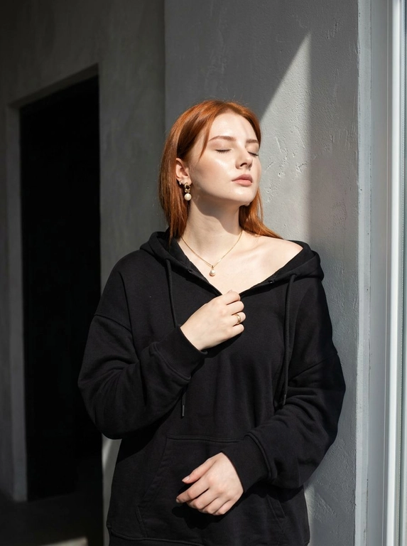 Используй загруженное фото для лица, строго не меняя черты лица, пропорции, возраст, текстуру кожи и волосы. Минималистичный портрет в интерьере с естественным светом, женщина стоит у серой текстурированной оштукатуренной стены, голова слегка запрокинута назад, глаза закрыты, лицо повернуто к источнику света, правая рука держит ткань толстовки у груди, левая рука расслабленно опущена вдоль тела. Оверсайз толстовка с капюшоном спущена с правого плеча, видны завязки, на шее тонкая золотая цепочка с кулоном с жемчужиной, золотые серьги среднего размера с жемчугом, золотое кольцо на пальце. Естественный макияж с деликатным скульптурированием, влажное свечение кожи, кожа ультрареалистичная без эффекта пластика и чрезмерной ретуши. Жёсткий направленный солнечный свет из окна создаёт резкие графичные тени на стене и лице, контрастное освещение, слева тёмный дверной проём добавляет глубину пространству. Съёмка на 50mm f/2.0, малая глубина резкости, естественный оконный свет, выраженные тени, цветовая гамма бежевый, золото, серый, баланс тёплых и холодных оттенков, minimalist editorial, фотореализм высокого качества.

Какой цвет толстовки?: Цвет толстовки чёрный.