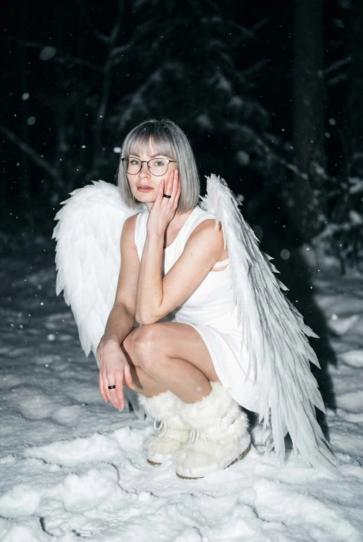  Создай фото человека на основе загруженной фотографии.
Белое платье без рукавов, пушистые белые сапоги, большие белые крылья, опущенные вниз, шире плеч.
Сидит на корточках на снегу, рукой касается щеки. На заднем плане заснеженный лес.
Ночная улица, тёмный размытый фон, вспышка.
Мягкое нечеткое фото: шум, зерно, лёгкое размытие, холодная атмосфера.
