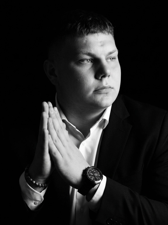 A hyper-realistic 8K cinematic portrait of a uploaded person,
captured in a dramatic Chiaroscuro style. The subject is seated at a
three-quarter angle, leaning slightly forward with a relaxed yet
commanding posture. His face is turned away from the lens, with
one side of his face clearly illuminated while the other is
completely lost in deep, velvety black shadows. His expression is
contemplative, not looking at the camera. The pose is precise: his
hands are clasped together near his chest; one hand is held
straight with fingers slightly tilted, while the other hand is
positioned so only the fingers are visible, resting over the first. He
wears a luxury black chronograph watch with a detailed metal link
bracelet, and a beaded bracelet. His attire is a sharp
black suit jacket over a white dress shirt with the top buttons
open, showing refined fabric textures. The background is a solid,
seamless black. The lighting is sharp and directional, highlighting
the intricate details of his hair,
the moisture in his eyes, and the glint off the watch face.
Cinematic black-and-white color grade with rich contrast and
smooth, natural skin transitions. Shot on 85mm lens, f/1.8, high-
resolution commercial photography