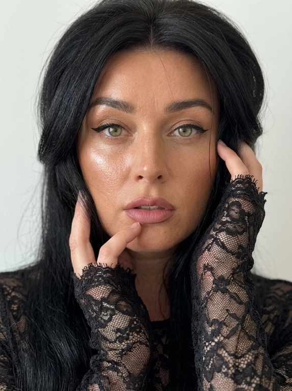Лёгкий макияж с акцентом на глаза: чёрные cat-eye стрелки на верхнем веке, нюдовая матовая помада оттенка пыльной розы, очерченная карандашом для губ на тон темнее, влажный хайлайтер. Кожа загорелая. Ультрареалистичная фотография, естественная текстура кожи, лёгкий шум, необработанный реалистичный вид, без фильтра красоты, без CGI, visible eye capillaries, natural skin texture with pores and micro-imperfections, depth of field, subsurface scattering, soft morning light, Tyndall effect, hyper-detailed iris, unedited RAW photo, 8K resolution. Максимально фотореалистичный студийный портрет, вертикальный формат, снято на iPhone 17 Pro, 8K.

Женщина сидит, но кадр настолько близкий, что место, на котором она сидит, бёдра и ноги полностью остаются за пределами кадра. РАКУРС ОЧЕНЬ БЛИЗКИЙ: камера прямо перед женщиной, на уровне глаз, объектив близко к лицу. В кадре только голова, плечи, руки и небольшая часть верхней части корпуса. Макушка близко к верхней границе кадра. Лицо почти фронтально, совсем немного повёрнуто в сторону, взгляд прямо в объектив, губы слегка приоткрыты. Женщина сидит расслабленно, корпус слегка подан вперёд.

Обе руки подняты к лицу очень компактно. Локти находятся близко друг к другу перед корпусом и направлены вниз, не разведены в стороны. Одна кисть поднята снизу к подбородку и губам, пальцы мягко согнуты, указательный палец касается нижней губы. Вторая кисть находится немного выше, возле противоположной щеки. Запястья практически рядом. Руки располагаются по обе стороны лица, возле щёк и висков.

На женщине тонкая чёрная полупрозрачная кружевная кофта по фигуре с высоким облегающим воротником. Всё полотно состоит из нежного ажурного кружева с мелким органичным растительным рисунком из тонких переплетающихся стеблей, небольших цветов и листьев, между которыми просвечивает кожа. Это цельное тонкое кружевное полотно, не сетка с нашитой вышивкой и не тяжёлый гипюр. Рукава экстремально длинные, закрывают запястья и почти полностью заходят на кисти до оснований пальцев. Края рукавов необработанные: кружево заканчивается непосредственно по своему рисунку, образуя тонкие ажурные выступы вокруг кистей, без манжет, подгиба и ровной строчки.

Макияж выразительный: загорелая, увлажнённая, сияющая кожа с натуральной текстурой и порами, сильный бронзер и контуринг скул, влажный хайлайтер, вытянутые чёрные кошачьи стрелки, пухлые нюдово-розовые губы с более тёмным коричнево-розовым контуром.

Позади неё ровная однотонная молочная стена: светлый мягкий молочно-белый оттенок, не серая, не серо-молочная, не бежевая, без декора и предметов. Мягкий нейтральный студийный свет, натуральный баланс белого, естественные оттенки кожи. Никакой жёлтой, золотистой или тёплой обработки. Чистая натуральная фотография без фильтров, пресетов, цветокоррекции, винтажного эффекта, HDR, ретуши, боке, пластиковой кожи и ИИ-эффекта. Маникюр молочный, миндальной формы, ровный и аккуратный.

Причёска: Волосы с сильным воздушным объёмом у корней, уложены на крупный брашинг. Передние объёмные слои проходят вдоль висков и щёк, слегка приминаются поднятыми руками и ниже снова становятся пышными. Несколько тонких прядей естественно проходят возле щёк и губ. Никакой расчёски, щётки или других предметов в кадре.