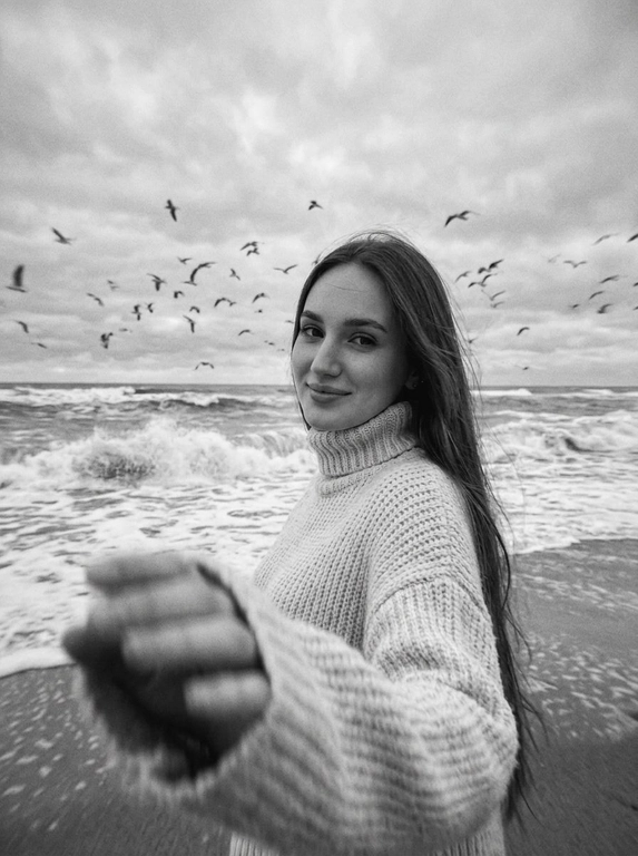Создай портрет, не меняя черты лица. Черно-белая фотография в стиле ретро: человек стоит на морском побережье, в полупрофиль, смотрит нежно в камеру, легкая улыбка на лице, одет(а) в светлый оверсайз вязаный свитер с высоким воротником. На переднем плане — размытая рука в движении (motion blur), на заднем плане — море с волнами и стая птиц в небе, пасмурное небо. Сильная зернистость пленки, винтажная эстетика, атмосфера свободы, эффект размытия в движении, эстетика.