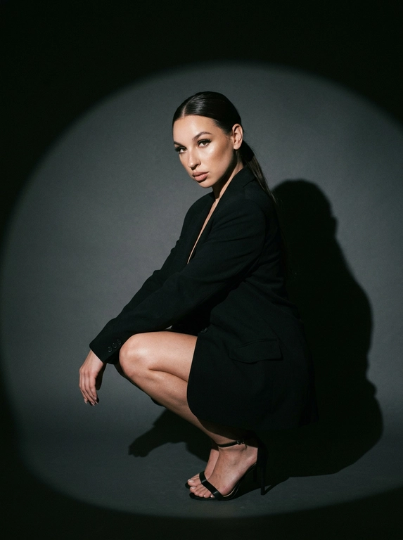 Модная студийная съёмка в стиле fashion editorial. Не менять внешность женщины. Женщина одета в только в чёрный оверсайз пиджак. Профессиональный макияж с акцентом на кожу и глаза. Один направленный прожектор формирует чёткое круглое световое пятно на темно-сером бесшовном гладком фоне и выразительную тень за спиной. Контрастный кинематографичный свет, глубокие тени, драматичный минимализм. Атмосфера — уверенность, сила, эстетика high fashion. высокая детализация кожи, реалистичная текстура, съёмка уровня модного журнала. Средний план. Женщина сидит на корточках, одна рука лежит на колене, корпус слегка наклонён вперёд. Уверенный прямой взгляд в камеру. На ногах чёрные босоножки на тонком каблуке. Чёткая тень позади усиливает драматизм кадра.

Какая будет прическа?: Волосы уложены в гладкий хвост.