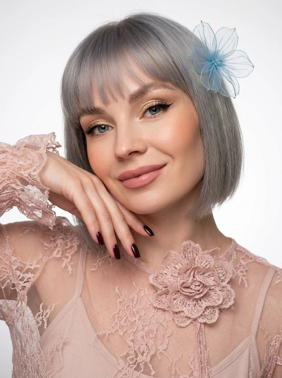 На фотографии запечатлена женщина крупным планом, позирующая для портрета. Снимок подчеркивает её ухоженную внешность и стильный образ.
Лицо и выражение:
•  Глаза:  ярко выделены макияжем. На верхних веках нанесены мерцающие золотисто-бежевые тени, дополненные четкими, длинными черными стрелками («кошачий глаз»). Ресницы густые и длинные, подчеркивающие глубину взгляда.
•  Брови: Густые, уложенные вверх (эффект ламинирования), что придает лицу современный и открытый вид.
•  Кожа: Тон кожи ровный, с нежным персиковым румянцем на скулах. Кончик носа и скулы слегка подсвечены хайлайтером для придания лицу сияния.
•  Губы: Выразительные, накрашены матовой помадой нежно-розового оттенка. Верхняя губа имеет четкий контур.
•  Поза: Женщина слегка наклонила голову вправо и подперла щеку правой рукой. Этот жест придает снимку нотку нежности и кокетства. Маникюр темно-бордовый,квадратный.
В волосах один нежный, воздушный цветок.
Одежда: На женщине надета кружевная блузка из легкой, полупрозрачной ткани.
•  Ткань украшена в крупный кружевной цветок .
•  Рукава имеют пышные воланы или оборки у запястий, что добавляет образу романтичности и женственности.
Фон и освещение:
•  Фон белый студийный
•  Освещение мягкое, направленное на лицо, что позволяет рассмотреть все детали макияжа и создает приятную, эстетичную картинку.
Общее впечатление:
Фотография представляет собой классический бьюти-портрет. Образ женщины сочетает в себе элегантность, современность и романтичность. Тщательно проработанный макияж и стильная одежда создают гармоничный и привлекательный облик.

Какого цвета будет блузка?: Цвет блузки нежно розовый.

Какого цвета будет цветок?: Цвет цветка в волосах синий.