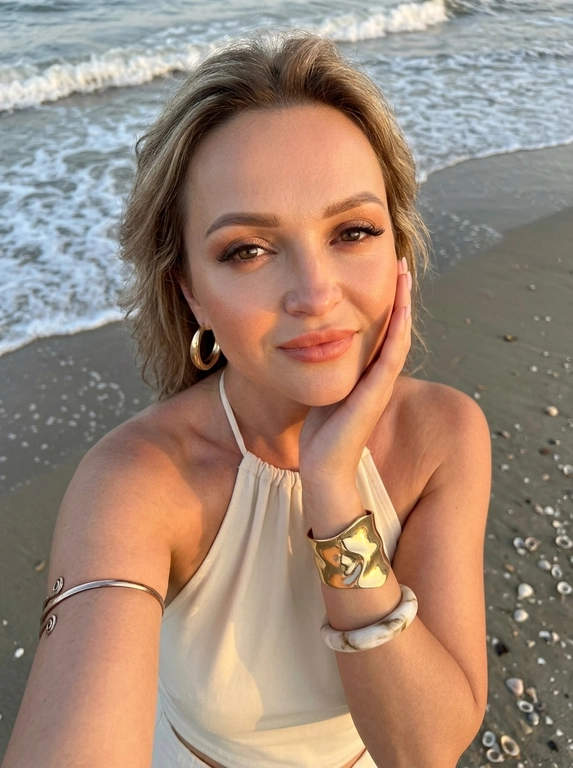 ultra realistic lifestyle beauty selfie photo of a woman standing on a sandy beach shoreline near the water, vertical composition shot from above with a smartphone held in one hand, camera angled downward creating a high perspective typical for handheld selfie photography, framing from chest upward. The woman slightly tilts her head upward toward the camera while her free hand gently touches the side of her face with fingers resting along the cheek and jawline, shoulders slightly angled creating a relaxed elegant pose. She is wearing a cream halter neck swimsuit or halter summer top with open shoulders and exposed collarbones. Accessories include large gold hoop earrings, a thick sculptural gold bracelet, a light marble-style bangle and a thin metallic arm bracelet placed above the elbow. Makeup soft glamorous beach style: luminous skin with warm sun glow, softly defined cheekbones with peach blush, clean sculpted brows, subtle winged eyeliner, long lashes and satin lips. Background shows wet beach sand with natural textures and a thin line of sea foam from a small wave approaching behind her, scattered small shells visible on the sand. Lighting natural sunset or golden hour coastal light coming from the side, warm highlights on skin and soft shadows around facial contours, shallow depth of field, realistic DSLR smartphone hybrid aesthetic, candid summer vacation mood