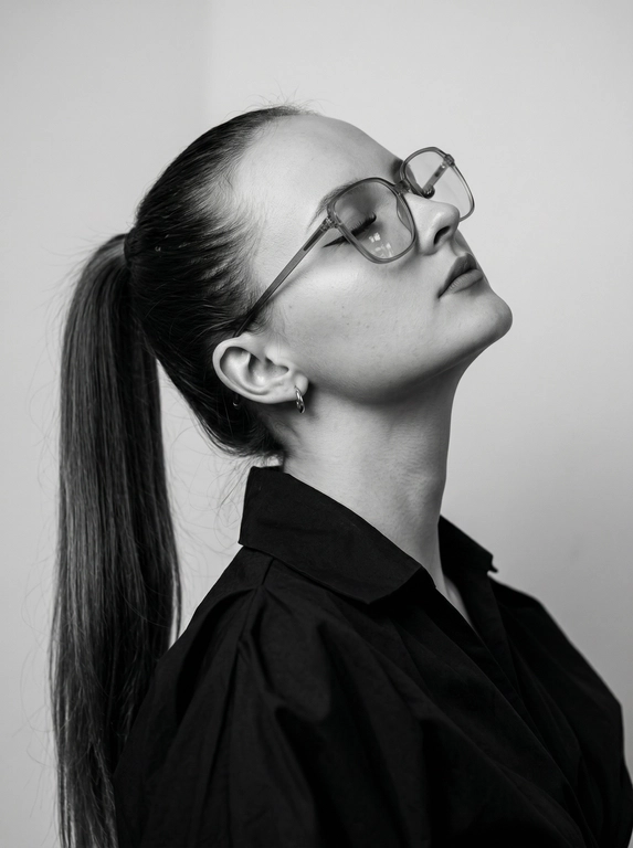 Чёрно-белый минималистичный фотореалистичный портрет, студийный мягкий свет спереди‑сверху, нейтральная светлая однотонная стена как фон. Девушка в профиль: голова запрокинута назад, подбородок поднят, глаза закрыты — расслабленная fashion-поза с акцентом на линии шеи и контуры лица. Обязательно точно сохранить черты лица с референс-фото (без искажений); крупные квадратные очки в массивной полу-прозрачной оправе с естественными бликами. Детали лица: длинные ресницы и чёткие стрелки, выразительные губы, естественная текстура кожи, аккуратный ретуш. Одежда: тёмный объёмный верх с выразительным воротником, маленькая серьга-кольцо. Стиль: высокая графичность и контраст, чистые линии, лёгкая драматичность, максимальная детализация и фотореализм, аккуратные светотеневые переходы, подчёркнутые контуры.

Какая будет прическа?: Гладкие волосы, высокий длинный густой хвост.