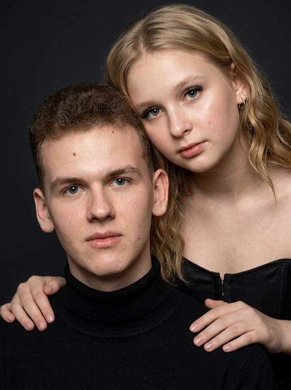 Гиперреалистичный студийный fashion-портрет молодой пары по загруженному референсу, 100% сходство лиц. Черты лица не изменять. Крупный план, кадр по плечи. Мужчина, спокойный уверенный взгляд прямо в камеру, естественное выражение лица, одет в черный трикотажный свитер с высоким воротом. Женщина стоит позади мужчины и нежно обнимает его, ее руки лежат на его плечах. Ее лицо расположено рядом с его лицом, она повернута слегка боком, голова немного наклонена вперед, взгляд направлен прямо в камеру. У женщины выразительные глаза с длинными густыми черными ресницами, естественный clean-glam макияж, натуральные брови, теплый оливковый оттенок кожи, небольшие серьги-кольца. Композиция плотная и интимная, их лица расположены очень близко друг к другу, современный модный портрет пары. Женщина одета в черный корсет. Кожа максимально натуральная: живая текстура кожи, видимые микропоры, subtle skin glow, естественный мягкий блеск кожи, natural skin microtexture, skin translucency, micro skin details, легкие естественные несовершенства кожи, без ретуши и без эффекта пластика. Освещение: мягкий студийный beauty lighting, рассеянный ключевой свет спереди и немного сбоку, мягкие глубокие тени для объема лица, деликатные световые блики на коже. Фон: глубокий темный студийный фон, темно-графитовый или почти черный, создающий контраст с кожей и лицами. Стиль: luxury fashion editorial photography, high fashion portrait, ultra photorealistic, 85mm portrait lens, shallow depth of field, creamy bokeh, cinematic soft lighting, RAW photo, extremely detailed, fine hair strands, natural skin pores, professional photography, photorealism, 8k, high dynamic range.