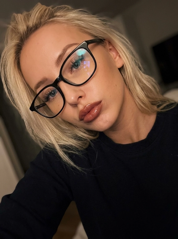 Селфи молодой женщины крупным планом, с высоким ракурсом, в мягкой, теплой эстетике. У нее очень длинные светлые волосы с небрежно уложенные вокруг лица, и она носит большие очки в черной оправе. Губы у нее средней полноты блестящие, с 
Коричнево-розовой помадой, слегка надутые. Ее макияж глаз включает в себя яркую подводку для глаз и длинные черные ресницы. На ней темный, возможно, черный топ с длинными рукавами. Композиция представляет собой диагональный портрет с наклоненной головой, занимающей большую часть кадра. Фон мягко размыт и темен, что предполагает обстановку в помещении с низким рассеянным освещением, возможно, от одного источника света сверху или сбоку, что создает мягкие тени и блики на ее коже и волосах. Общее настроение интимное и расслабленное. Освещение мягкое и теплое, подчеркивающее ее черты лица, особенно губы и отражения в очках. linapromts В цветах преобладают теплые тона кожи: кораллово-розовый для губ, темно-черный для одежды и очков, с нежными коричневыми и светлыми оттенками волос на приглушенном темном фоне. Стиль — современная селфи-фотография, слегка мечтательная или стилизованная. Эффект живого кадра, мягкий цифровой шум, немного зернистости, фото в движении. Черты лица сохранены как на исходном фото. Фото 2:3