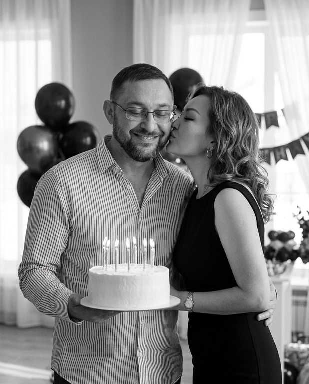 Create an ultra-photorealistic, cinematic black-and-white birthday portrait of a young romantic couple.
A man with, wearing a thin vertically striped button-up shirt, smiling naturally with his eyes closed. He holds a round white-frosted birthday cake with six lit candles at waist level.
A woman stands closely on his right, gently kissing his cheek with her eyes closed. wears an elegant fitted black sleeveless dress with a modest neckline and a delicate wristwatch. Her arm wraps naturally around his waist, while his arm embraces her.
Elegant indoor birthday setting with clusters of black balloons, large windows, sheer white curtains, and soft natural daylight. Intimate, romantic, spontaneous atmosphere.
Professional 85mm portrait photography, realistic skin pores, natural hair strands, authentic anatomy, soft cinematic lighting, rich monochrome contrast, subtle film grain, realistic candle flames, 8K photographic quality.
Negative: CGI, plastic skin, excessive smoothing, distorted faces, unnatural hands, extra fingers, artificial expressions, cartoon, beauty filters, unrealistic anatomy.