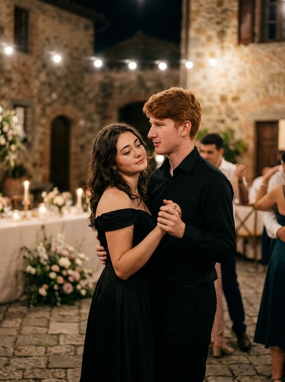 Cinematic realistic photo of a man and a woman slow dancing intimately at night. The man is wearing a black button-up shirt. The woman is wearing an elegant black off-the-shoulder evening dress. They are dancing closely together at an outdoor wedding venue in an old Italian village courtyard. The mood is romantic, tender and intimate. Warm dim lighting comes from distant string lights and candles, creating soft highlights and gentle shadows. In the softly blurred background, wedding tables, decor and a few guests are visible, adding depth and atmosphere without distracting from the couple. Their body language feels natural and emotional, with a deep affectionate look between them. The image should feel like a real candid moment captured from the side by someone nearby, not a staged fashion shoot. Shot on iPhone, ultra-realistic, natural skin texture, realistic fabric details, slight low-light grain, soft background focus, authentic evening atmosphere, high detail.
