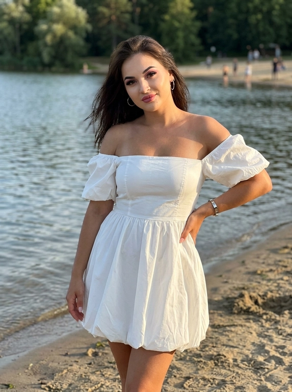 Фотореалистичная lifestyle-фотография в вертикальном формате 4:5, снятая на iPhone 17 Pro Max, объектив 1×. Женщина стоит у кромки спокойной реки или озера на песчаном пляже тёплым летним вечером.

Камера расположена немного ниже уровня груди. Кадр имеет лёгкий естественный наклон. Женщина показана от головы примерно до колен, чтобы хорошо читались фигура и поза. Она слегка наклоняет корпус в сторону камеры, голова также немного наклонена. Взгляд направлен прямо в объектив, на лице лёгкая уверенная полуулыбка.

Волосы слегка развеваются тёплым вечерним ветром. Кожа ровная, загорелая, с естественным сатиновым сиянием. Макияж soft glam: ровный тон, лёгкий бронзер, тонкие стрелки, длинные ресницы, аккуратно оформленные брови и губы с мягким сатиновым финишем.

Правая рука лежит на бедре, левая свободно опущена вдоль тела. Поза расслабленная, естественная и уверенная. Ноги видны примерно до уровня колен.

На заднем плане спокойная голубая вода с лёгкой рябью, удалённая береговая линия с зелёными деревьями и несколько отдыхающих далеко на фоне.

Освещение тёплого золотого часа. Мягкий солнечный свет падает сбоку, освещает лицо и кожу, создаёт естественный объём, деликатные блики и длинные мягкие тени.

Дорогая летняя lifestyle-съёмка, тёплые золотистые оттенки, насыщенный синий цвет воды, натуральные тона кожи, слегка пониженная экспозиция, мягкий контраст, лёгкая цифровая зернистость, реалистичный эффект мобильной камеры iPhone, RAW, максимальный фотореализм, высокая детализация, атмосфера дорогого летнего отдыха, quiet luxury, natural candid elegance, без AI-глянца, без текста и водяных знаков.

Какая будет одежда?: На женщине белое короткое платье-баллон из плотной матовой тафты с открытыми плечами, чётко сформированным лифом и объёмной округлой юбкой чуть выше колен. Скульптурная форма ткани красиво держится в кадре и контрастирует со спокойным природным фоном. Образ дополнен тонкими серьгами и гладким жёстким браслетом. Выглядит современно, дорого и заметно отличается от обычных пляжных комплектов.