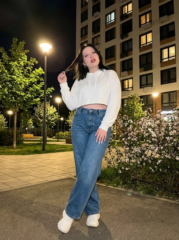 Не меняя внешность. Фото снято ночью на iPhone. Женщина в городском дворе, мягкий тёплый свет фонаря, вокруг зелень, цветущие деревья и современный жилой комплекс.
Она стоит расслабленно, слегка развернувшись в сторону, ноги перекрещены. Одна рука играет с прядью волос. Взгляд — кокетливый, уверенный.
Образ современный и молодёжный: укороченная худи, широкие джинсы с высокой посадкой и массивные кроссовки. Минималистичные украшения — тонкие серьги-кольца.
Макияж аккуратный: ровный сияющий тон, графичные брови, длинные ресницы с «лисий» эффектом, розово-ягодные губы с блеском, лёгкий хайлайтер на скулах. Ногти — молочный миндаль.
Съёмка со вспышкой: лёгкое зерно, мягкая резкость, высокая детализация кожи и ткани, эффект живого кадра.

Цвет худи: Цвет худи белый