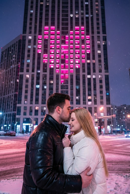   Создай не меняя черты лица. Романтический снимок пары, стоящей на заснеженной городской улице поздним вечером. Главный объект – крупный план пары: мужчина в черной кожаной дутой зимней куртке нежно целует женщину в белой дутой куртке в лоб, она стоит с прикрытыми глазами, улыбаясь. За ними возвышается массивный современный жилой небоскреб, на фасаде которого окна на нескольких этажах подсвечены ярким  светом, формируя огромное пиксельное сердце. Освещение драматичное, контрастное, с преобладанием холодных тонов в небе и тёплого, неонового розового свечения от здания. Снег на земле отражает свет. Урбанистическая, зимняя, кинематографическая фотография, атмосферный кадр.
