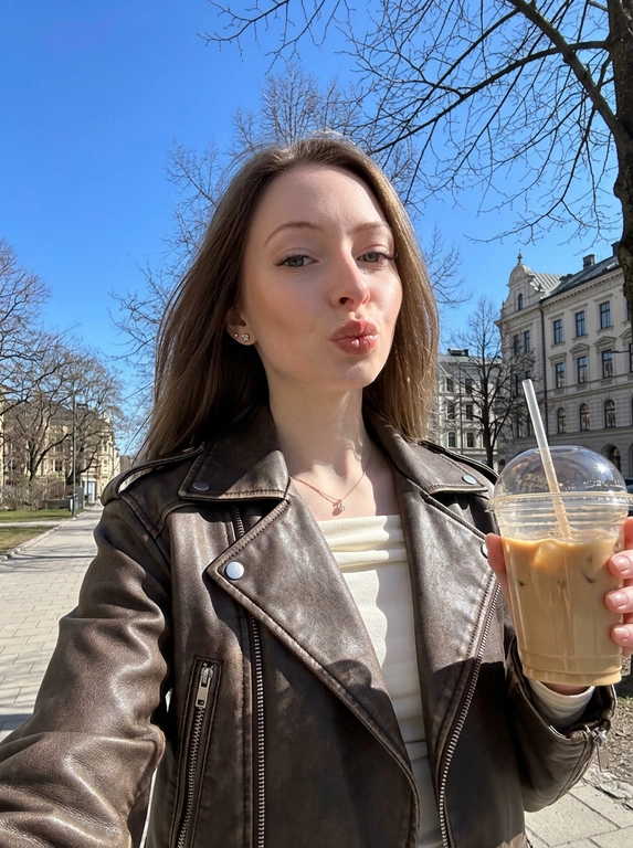Не меняй черты лица женщины с фото. На этом ярком солнечном снимке запечатлена женщина в эстетике весенней прогулки. Фотография выполнена в формате селфи на фоне ясного голубого неба и деревьев. У неё аккуратный макияж с акцентом на сияющие губы (блеск), чётко оформленные брови, нарощенные ресницы в «лисьем» эффекте, лёгкий контуринг на скулах. На ней стильная кожаная косуха шоколадного цвета с эффектом состаренности. Под курткой виден лонгслив из вискозы с открытыми плечами и напуском молочного цвета. В ушах видны серьги-гвоздики и тонкая цепочка на шее с маленьким бриллиантом. Яркий прямой солнечный свет создаёт чёткие контрасты и подчёркивает текстуру одежды и блеск аксессуаров. Фон: чистое ярко-голубое небо без единого облака, ветви деревьев без листвы, асфальтовый тротуар, вдали красивые архитектурные здания, женщина прогуливается по улице города. Общее впечатление — позитивное прогулочное настроение. Ракурс: на вытянутой руке, снизу вверх.

Кофе в руке:  В руке пластиковый стакан кофе с трубочкой.