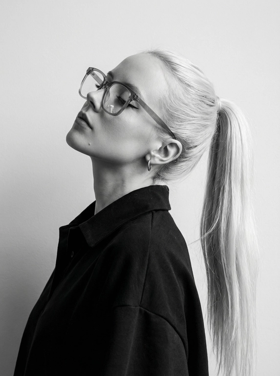 Чёрно-белый минималистичный фотореалистичный портрет, студийный мягкий свет спереди‑сверху, нейтральная светлая однотонная стена как фон. Девушка в профиль: голова запрокинута назад, подбородок поднят, глаза закрыты — расслабленная fashion-поза с акцентом на линии шеи и контуры лица. Обязательно точно сохранить черты лица с референс-фото (без искажений); крупные квадратные очки в массивной полу-прозрачной оправе с естественными бликами. Детали лица: длинные ресницы и чёткие стрелки, выразительные губы, естественная текстура кожи, аккуратный ретуш. Одежда: тёмный объёмный верх с выразительным воротником, маленькая серьга-кольцо. Стиль: высокая графичность и контраст, чистые линии, лёгкая драматичность, максимальная детализация и фотореализм, аккуратные светотеневые переходы, подчёркнутые контуры.

Какая будет прическа?: Гладкие волосы, высокий длинный густой хвост.