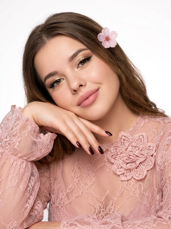 На фотографии запечатлена женщина крупным планом, позирующая для портрета. Снимок подчеркивает её ухоженную внешность и стильный образ.
Лицо и выражение:
•  Глаза:  ярко выделены макияжем. На верхних веках нанесены мерцающие золотисто-бежевые тени, дополненные четкими, длинными черными стрелками («кошачий глаз»). Ресницы густые и длинные, подчеркивающие глубину взгляда.
•  Брови: Густые, уложенные вверх (эффект ламинирования), что придает лицу современный и открытый вид.
•  Кожа: Тон кожи ровный, с нежным персиковым румянцем на скулах. Кончик носа и скулы слегка подсвечены хайлайтером для придания лицу сияния.
•  Губы: Выразительные, накрашены матовой помадой нежно-розового оттенка. Верхняя губа имеет четкий контур.
•  Поза: Женщина слегка наклонила голову вправо и подперла щеку правой рукой. Этот жест придает снимку нотку нежности и кокетства. Маникюр темно-бордовый,квадратный.
В волосах один нежный, воздушный цветок.
Одежда: На женщине надета кружевная блузка из легкой, полупрозрачной ткани.
•  Ткань украшена в крупный кружевной цветок .
•  Рукава имеют пышные воланы или оборки у запястий, что добавляет образу романтичности и женственности.
Фон и освещение:
•  Фон белый студийный
•  Освещение мягкое, направленное на лицо, что позволяет рассмотреть все детали макияжа и создает приятную, эстетичную картинку.
Общее впечатление:
Фотография представляет собой классический бьюти-портрет. Образ женщины сочетает в себе элегантность, современность и романтичность. Тщательно проработанный макияж и стильная одежда создают гармоничный и привлекательный облик.

Какого цвета будет блузка?: Цвет блузки нежно розовый.

Какого цвета будет цветок?: Цвет цветка в волосах розовый.