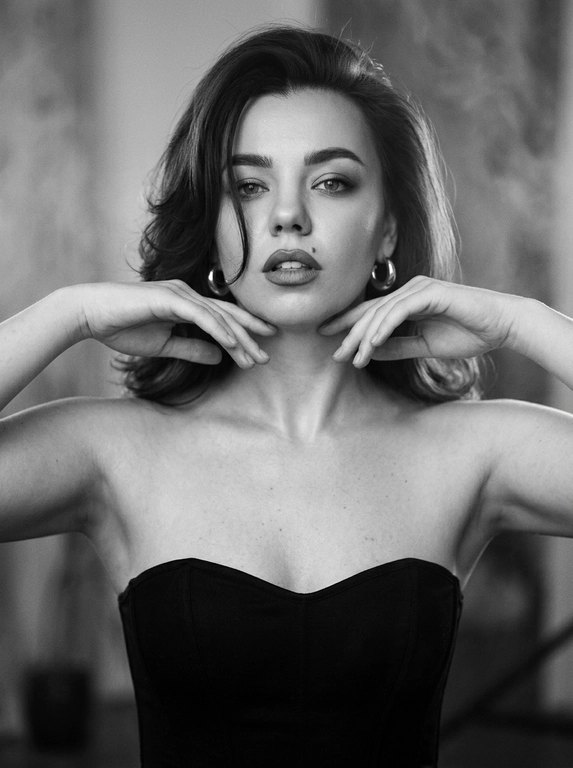 Кинематографический эффект с добавлением зерна. 
Вертикальный fashion-портрет, чёрно-белая плёночная съёмка в стиле editorial. 
Кадр по пояс, лёгкий поворот корпуса, плечи по диагонали, взгляд в камеру. Техника - 85mm, Kodak Tri-X 400, мягкая тональность, сильное зерно, легкий шум и немного дыма. Драматичные тени. Безупречный макияж, губы с более тёмным контуром, слегка приоткрыты. Лёгкое движение головы и корпуса с деликатным motion blur,  подбородок приподнят, руки у лица.
В ушах толстые серьги кольца.
Чёрный корсетный топ без бретелей, акцент на плечах и шее. Мягкий студийный свет спереди и сбоку, плавные тени. Ультрареализм, натуральная текстура кожи, monochrome film editorial aesthetic.

Какая будет укладка? : Волосы каскадом, брашинг с объёмом у корней, концы подкручены внутрь, одна прядь падает на лицо.