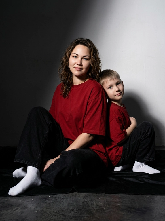 Гиперреалистичная фотография. Качество 8К. Используя портретное сходство с людьми с фотографий, создай фотографию: женщина в красной оверсайз свободной футболке и свободных черных джинсах, белых носках. Ребенок одет в красную оверсайз футболку, черные свободные джинсы и белые носочки. Женщина сидит на полу, согнув колени, смотрит прямо в кадр с еле заметной улыбкой. Сзади женщины в такой же позе сидит ребенок облокотившись спиной к спине женщины, с легкой улыбкой. Темный стиль модного редакционного журнала, глубокие тени и драматическое студийное освещение, белый фон, элегантная и мощная композиция, ультрареалистичный, высокая детализация, художественный портрет.