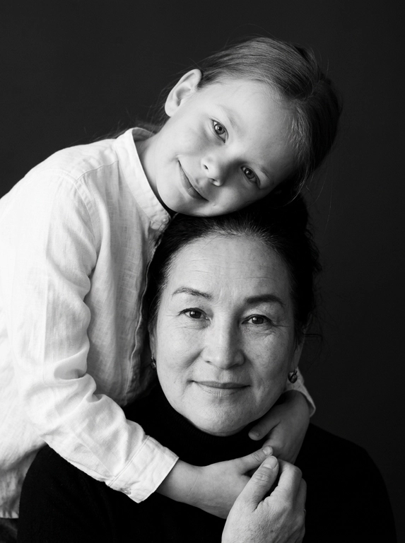Родитель и ребёнок с внешностью как на приложенных фото, художественная семейная студийная фотосессия. Композиция — вертикальный портрет, крупный план лиц. Родитель внизу смотрит прямо в камеру, лицо фронтально. Ребёнок сверху, голова повернута набок, щека лежит на голове родителя, смотрит в камеру с нежной улыбкой. Рука ребёнка обнимает родителя за шею, видна в кадре, родитель держит руку ребёнка своей рукой.
Одежда: ребёнок в белой льняной рубашке, родитель в чёрной водолазке. 
Фон — тёмно-серый почти чёрный однотонный студийный, создаёт контраст. Освещение — мягкое боковое студийное, создаёт объём и глубину.
Чёрно-белая художественная фотография с выразительными контрастами. Атмосфера нежная, интимная, спокойная.