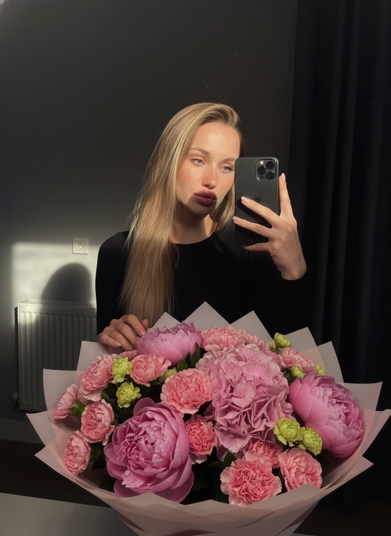 На фото — интерьерная зеркальная селфи-композиция на айфон 17 pro. Центральный объект в кадре —девушка в чёрной рубашке. Волосы длинные и прямые, падают ниже плеч, на свету блестят и дают тёплые блики.Макияж нюд, слегка загорелая кожа,, длинные ресницы, блеск на губах. Сходство 100%.Девушка держит телефон в правой руке, аппарат в тёмном (чёрном) чехле с модулем камеры в верхнем углу: заметна круглая конфигурация объективов. Левой рукой она опирается на поверхность стола или пола позади большого букета. Ногти аккуратно накрашены в светлый оттенок. На переднем плане очень крупный пышный букет в розовой цветовой гамме, упакованный в бумагу пастельных тонов. В букете разнообразные цветы: крупные махровые пионы/гвоздики/гортензии или похожие по форме рuffled-бутоны разной интенсивности розового, а также более мелкие бутончики светло-зелёного и кремового оттенков, которые создают цветовые акценты и объём. Композиция плотная, насыщенная, выглядит свежей и профессионально оформленной.Освещение контрастное — естественный солнечный луч слева создаёт яркие пятна и тёплые блики на волосах и букете, при этом образ окружён тенью, что даёт драматический эффект и подчёркивает фактуру цветов. Фон минималистичен: нейтральная тёмно-серая стена, с правой стороны видна тёмная штора, слева — часть панельного радиатора и неглубокий прямоугольный предмет (возможно элемент декора или обогревателя). На стене заметна электрическая розетка. Композиция кадра подчёркивает букет и силуэт, использует контраст света и тени для создания атмосферного, интимного настроения.