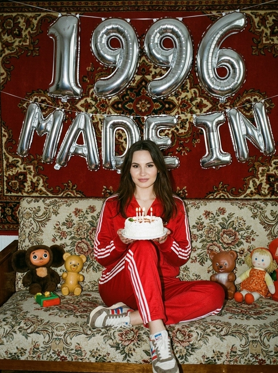Создай крупный портрет, не меняя черты лица. На фотографии модель в стиле «лихие 90-е» в России. Модель сидит на старом диване с цветочной обивкой на фоне красного советского ковра на стене и смотрит в камеру с лёгкой улыбкой. На ковре висят большие надувные серебряные фольгированные шары в форме цифр и надпись «MADE IN». На модели классический красный спортивный костюм того времени и кроссовки. В руках маленький праздничный белый торт со свечами. Рядом на диване лежат игрушки: советские игрушки и старая советская кукла. Фото со вспышкой. Освещение мягкое, создающее атмосферу ностальгии. Высокое разрешение, реализм. Модель в олдскульном спортивном костюме сидит расслабленно, с лёгкой улыбкой. Ретро-вайб, приглушённые цвета, зернистость плёнки, детализированные текстуры ткани и шерстяного ковра.

Какая будет цифра?: Цифра 19966.