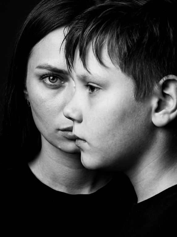 Чёрно-белый фотореалистичный студийный beauty-fashion портрет родителя и ребёнка с представленных фото. Крупный плотный кадр по плечи, без свободного пространства. Ребёнок находится на переднем плане, ближе к камере, в профиль, расположен сбоку и частично перекрывает лицо родителя. Лицо ребёнка закрывает примерно половину лица родителя, создавая сильный эффект визуального перекрытия. Родитель находится позади ребёнка, строго по центру кадра; у родителя виден только один глаз, направленный прямо в камеру, вторая часть лица скрыта лицом ребёнка. Лица расположены максимально близко, почти соприкасаются, но без явного физического действия. Ребёнок смотрит в сторону, выражение спокойное, сдержанное и закрытое. Родитель смотрит прямо в камеру одним открытым глазом, взгляд холодный, спокойный и пронизывающий. Атмосфера high-fashion editorial: минимальная эмоциональность, люксовая отстранённость, скрытое напряжение и холодная близость. Фон тёмный, чистый, студийный, без деталей. Жёсткое студийное освещение, высокий контраст, глубокие тени, выразительная светотень на лицах, натуральная текстура кожи, sharp focus, dramatic black and white photography, editorial beauty portrait, без улыбок, без лишних предметов, без текста и логотипов.