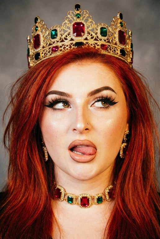 Создай фото -детальное описание художественного портрета девушки, выполненного в популярной современной эстетике, сочетающей элементы «е-qirl» и стиля «rebellious royalty» (бунтарская королевская особа).
:Центральный образ и внешность:
• Лицо и поза: Девушка позирует в дерзкой и провокационной манере.
Её глаза закатаны (или направлены сильно в сторону и вверх), а кончик языка слегка высунут. Это создает образ, сочетающий в себе игривость, бунт и некоторую отрешенность.
Элегантный макияж глаза с длинными острыми черными стрелками, длинные густые ресницы эффект лучики, нижние ресницы накрашены, хайлайтер во внутренних уголках и скулах.
Пухлые полностью прокрашенные телесной матовые губы слегка приоткрыты легкий блеск и светло-коричневый карандаш по краям. персиково-розового оттенка.
Не меняй мою прическу. 
Корона: На голове массивная высокая корона в винтажном стиле. Она украшена сложным филигранным узором и крупными драгоценными камнями: по центру расположен большой квадратный красный камень (рубин), по бокам - камни зеленого (изумруд) и черного цветов. Корона сидит немного небрежно, подчеркивая неформальность образа.
Ожерелья:
На шее надет чокер, выполненный в одном стиле с короной - цепочка с круглыми красными камнями в оправе. В ушах серьги по такому же типо как корона •
Фон: Нейтральный, дымчато-серый или слегка туманный. Он лишен деталей, что позволяет полностью сфокусировать внимание на ярких аксессуарах и лице модели.
Освещение и обработка: Свет мягкий, рассеянный. Фотография имеет характерную обработку с эффектом «зернистости» (пленочное зерно), что придает ей винтажный и художественный вид.
Цветовая гамма теплая, с акцентом на золото и красный цвет.
Общее впечатление:
Снимок транслирует идею современной интерпретации власти и красоты. Это противопоставление классического символа величия (короны) и современного неформального поведения.
Образ выглядит стильно и кинематографично и запоминающе, сильная вспышка. Корона цвета золота. Глаза карие 