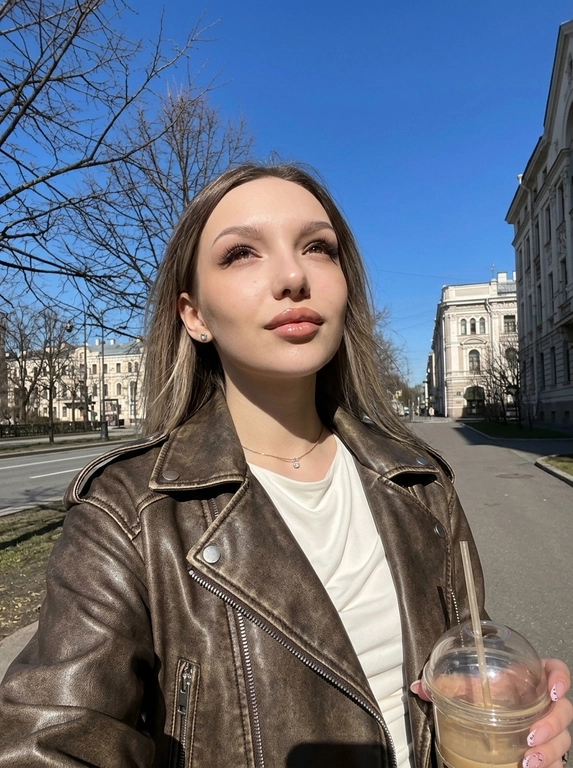 Не меняй черты лица женщины с фото. На этом ярком солнечном снимке запечатлена женщина в эстетике весенней прогулки. Фотография выполнена в формате селфи на фоне ясного голубого неба и деревьев. У неё аккуратный макияж с акцентом на сияющие губы (блеск), чётко оформленные брови, нарощенные ресницы в «лисьем» эффекте, лёгкий контуринг на скулах. На ней стильная кожаная косуха шоколадного цвета с эффектом состаренности. Под курткой виден лонгслив из вискозы с открытыми плечами и напуском молочного цвета. В ушах видны серьги-гвоздики и тонкая цепочка на шее с маленьким бриллиантом. Яркий прямой солнечный свет создаёт чёткие контрасты и подчёркивает текстуру одежды и блеск аксессуаров. Фон: чистое ярко-голубое небо без единого облака, ветви деревьев без листвы, асфальтовый тротуар, вдали красивые архитектурные здания, женщина прогуливается по улице города. Общее впечатление — позитивное прогулочное настроение. Ракурс: на вытянутой руке, снизу вверх.

Кофе в руке?:  В руке пластиковый стакан кофе с трубочкой.