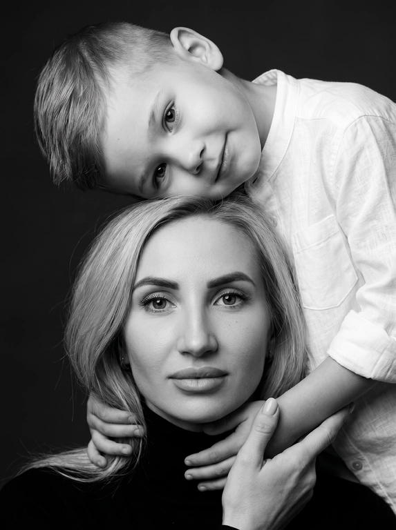 Родитель и ребёнок с внешностью как на приложенных фото, художественная семейная студийная фотосессия. Композиция — вертикальный портрет, крупный план лиц. Родитель внизу смотрит прямо в камеру, лицо фронтально. Ребёнок сверху, голова повернута набок, щека лежит на голове родителя, смотрит в камеру с нежной улыбкой. Рука ребёнка обнимает родителя за шею, видна в кадре, родитель держит руку ребёнка своей рукой.
Одежда: ребёнок в белой льняной рубашке, родитель в чёрной водолазке. 
Фон — тёмно-серый почти чёрный однотонный студийный, создаёт контраст. Освещение — мягкое боковое студийное, создаёт объём и глубину.
Чёрно-белая художественная фотография с выразительными контрастами. Атмосфера нежная, интимная, спокойная.