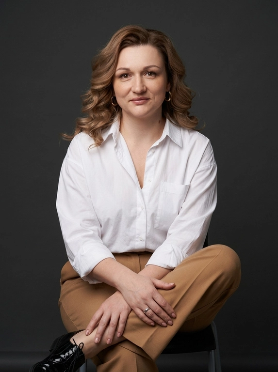 Создай фотореалистичный студийный editorial-портрет в стиле минималистичного глянцевого журнала. Образ — элегантный, уверенный, sophisticated, с атмосферой timeless elegance. 

Кадр — 3/4 роста, от макушки до середины голени. Камера расположена немного выше уровня глаз, с мягким естественным углом. Модель сидит на стуле с тонкими металлическими ножками темно-графитового цвета. Тело слегка развернуто вправо, голова повернута к камере. Корпус немного подан вперед, осанка уверенная и расслабленная. Ноги скрещены в элегантной позе, руки мягко сложены и лежат на верхней ноге. Взгляд прямой, уверенный, теплый, с едва заметной полуулыбкой.

На модели ее образ выглядит свежо, спокойно и элегантно.  Образ дополняют золотые серьги-кольца, тонкое серебряное кольцо. 

Макияж естественный и элегантный: ухоженные брови, легкая тушь, коричнево-бежевые тени, розовая помада, легкий румянец.

Фон — чистый студийный задник глубокого темно-серого антрацитового оттенка, с мягким градиентом и легкой рассеянной тенью от модели. Освещение профессиональное студийное: большой мягкий источник света слева спереди, мягкий заполняющий свет справа и легкий контровой свет сзади, подчеркивающий объем волос. Средний контраст, мягкие переходы теней, реалистичная детализация кожи, волос и ткани. Кадр должен выглядеть как дорогой editorial-портрет для модного журнала: чисто, минималистично, реалистично, без лишней ретуши.

Какая будет прическа?: Волосы объемные, мягко уложенные, с крупными свободными волнами и легкой живой текстурой. 

Выберите образ: Белая оверсайз-рубашка из хлопка с естественными складками, слегка расстегнутая у воротника, и широкие прямые брюки высокой посадки теплого кэмел-оттенка.Частично виден черный лакированный оксфорд.