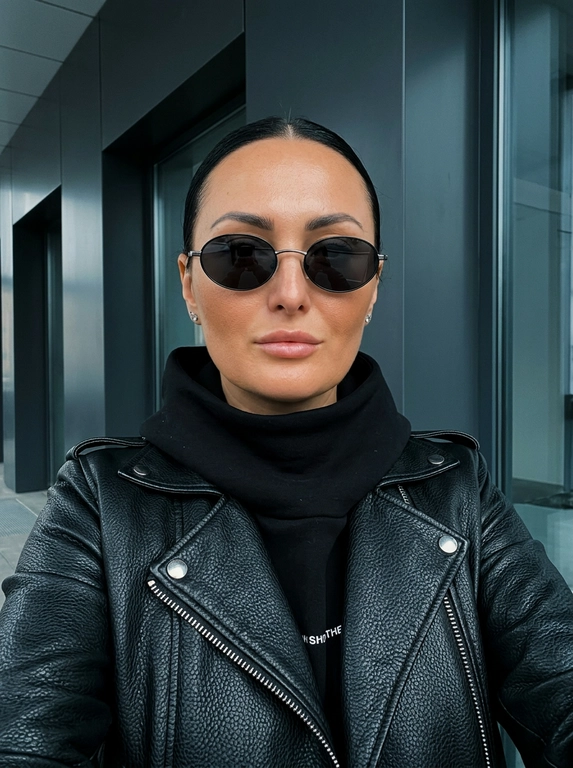Взгляд направленный прямо в объектив камеры.

Кожа: Ultra-realistic expensive-looking skin с видимыми порами, мягким персиковым пушком, реалистичной текстурой, тонкими тональными переходами, субповерхностным рассеиванием света и естественной асимметрией. Кожа должна выглядеть здоровой и профессионально ухоженной, как после премиальных косметологических процедур. Тёплый загорелый подтон с реалистичной глубиной и матово-сатиновым сиянием от холодного студийного света. Без чрезмерного сглаживания, пластиковой текстуры, CGI-идеальности и эффектов бьюти-фильтра. Кожа слегка загорелая.

Макияж: Минималистичный, но выразительный. 

Одежда: Стильный уличный образ. Чёрная кожаная объемная куртка из фактурной кожи (эффект "пеббл") с серебристой молнией и кнопками. Под курткой — чёрное худи с высоким объёмным воротником, закрывающим шею. На нижнем крае худи видна белая надпись (в стиле бренда).

Украшения и аксессуары: Крупные  серьги-гвоздики. 

Поза и действие: Расслабленная, уверенная поза. Съёмка снизу вверх (низкий ракурс), голова слегка наклонена вперёд, взгляд направлен прямо в камеру. Одна рука (за кадром) держит телефон для селфи, корпус слегка развёрнут.

Окружение: Современный минималистичный интерьер. Задний план — тёмные матовые архитектурные панели (чёрные или тёмно-серые) и стеклянное окно с отражающимся холодным светом. Атмосфера строгая, индустриальная и элегантная.

Освещение: Холодное, рассеянное естественное освещение. Свет создаёт глубокие, мягкие тени на лице и подчёркивает фактуру кожи и текстуру чёрной кожи куртки.

Техника: Sony A7 III, 85mm f/1.8, холодный студийный свет, чёткие детали, кинематографичный стиль, лёгкое зерно, RAW photo, высокая детализация. Не размывать фон! (Чёткие линии архитектурных панелей, стекла и фактура кожаной куртки должны быть идеально резкими, без размытия).

Ракурс: Низкий ракурс (снизу вверх) — камера расположена ниже уровня глаз, создавая эффект величия и уверенности.

План: Крупный план — кадр от груди до макушки, захватывающий лицо, очки, высокий воротник и фактуру кожаной куртки.

Какая будет прическа?: Волосы: Аккуратно зачёсаны назад и убраны в идеально гладкий тугой пучок. Никаких выбивающихся прядей, строгая и минималистичная укладка.

Очки: Чёрные овальные солнцезащитные очки с тонкой серебристой оправой.
