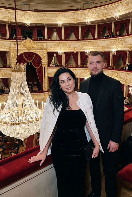 Я стою в ложе Большого театра, на рядов лож и большой хрустальной люстры. В ложах сидят люди, висят красные бархатные занавески. Камера на уровне глаз.  Съёмка выглядит как фото, сделанное на iPhone: естественный свет, лёгкое зерно, минимальная обработка, живой момент без позирования. Взгляд в камеру, легкая улыбка. Голова немного наклонена вбок. Волосы сияют, макияж естественный, губы глянцевые. Одна рука лежит на бортике ложа, во второй - маленькая черная сумочка. Приглушенный свет. На мне чёрное бархатное вечернее платье без рукавов по фигуре и белый жакет в стиле Channel. На мужчине черный пиджак и водолазка 