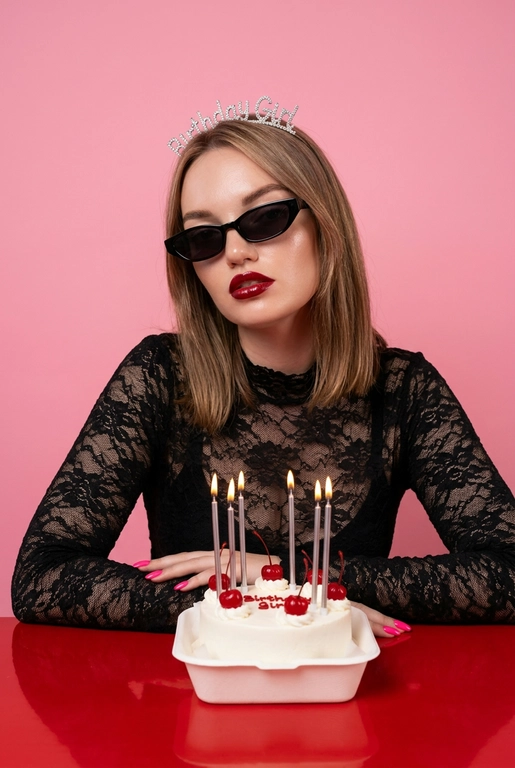Стильная fashion-фотосессия в студии с розовым фоном. Женщина сидит за красным столом, перед ней белый маленький бенто-торт с вишенками.  На торте тонкие серебристые зажженые свечки. Она в чёрном кружевном топе с длинными рукавами. На голове диадема с надписью “Birthday Girl”, на глазах чёрные узкие солнцезащитные очки. Макияж глянцевый, акцент на губах, чёткий контур, ухоженная кожа.
Женщина слегка наклонила голову в сторону, смотрит в камеру. Настроение кадра — дерзкий день рождения, fashion editorial, ironic glamour. Мягкий студийный свет, ровное освещение без жёстких теней, высокая детализация кожи, чистая композиция. Крупный план / средний план, фокус на лице, без искажений. Профессиональная фотосъёмка, модная журнальная эстетика.