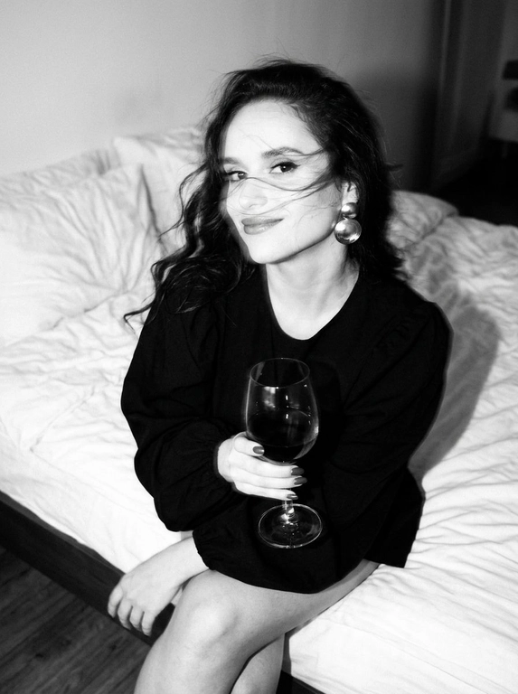 Create a portrait, transfer the face from the reference photo without changing facial features, proportions, or expression, preserve full identity. Aesthetic black-and-white nighttime Y2K photograph captured with direct on-camera flash, shot in motion.
A woman is sitting on the edge of a white, slightly rumpled bed, the frame taken from a slightly elevated angle. She wears a stylish black top with voluminous sleeves and large round statement earrings. In one hand she holds an elegant glass of dark wine. Her body is subtly moving, creating natural motion blur across the face and shoulders, as if the moment was caught mid-gesture. The gaze is languid and directed toward the camera, with a soft, intimate smile. Hair falls in large waves with loose strands drifting across the face, slightly displaced by movement. Lighting is harsh flash against deep night shadows, high contrast with blown highlights and strong falloff, visible film grain. Skin texture remains real and alive, no smoothing, enhanced by flash glare. The mood is high-fashion editorial, Parisian chic, old-money sensuality, nighttime melancholy, spontaneous and cinematic, shot in a raw 35mm film aesthetic with pronounced motion blur and lived-in realism.