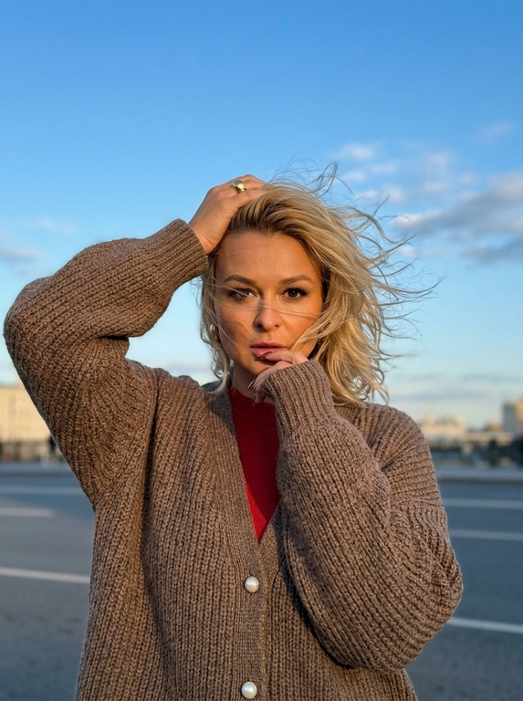 Сверхреалистичная уличная fashion-фотография на iPhone 17 Pro Max, основная камера 1x, 24 мм. Средний портрет примерно до середины бёдер, камера на уровне груди, женщина по центру кадра, над головой много открытого вечернего неба.

Женщина стоит на открытом пространстве, корпус почти фронтально к камере. Сильный ветер активно развевает волосы в стороны, отдельные тонкие пряди хаотично проходят перед лицом. Одна рука поднята над головой и лежит на макушке, локоть направлен в сторону; вторая рука согнута перед грудью, длинный рукав частично закрывает кисть, пальцы находятся возле губ. Взгляд прямо в объектив, губы сомкнуты, выражение уверенное, без улыбки.

На ней объёмный коричневый вязаный кардиган oversize с жемчужными пуговицами, длинными свободными рукавами и выраженной мягкой фактурой крупной вязки. Кардиган свободно собирается складками на теле.

Макияж выразительный, но естественный: кожа с лёгким загаром и натуральной текстурой, оформленные брови, длинные густые ресницы, мягкая чёрная подводка, лёгкий бронзер и румянец, пудрово-розовые губы, ногти коричневые.

Фон минималистичный: красивая вечерняя московская улица. Большую часть кадра занимает чистое голубое вечернее небо с небольшими мягкими светлыми облаками.

Тёплые золотистые блики на лице и волосах, мягкие естественные тени. Цветокоррекция слегка плёночная: приглушённые натуральные оттенки, тёплая кожа, немного пониженная экспозиция, умеренный контраст, лёгкая зернистость. Волосы должны двигаться физически естественно от сильного ветра. Без студийного света, пластиковой кожи, сильного боке, CGI и AI-эффекта. Кадр будто сделан в спешке и размыт на 2%.

Доп. характеристики: красный свитер