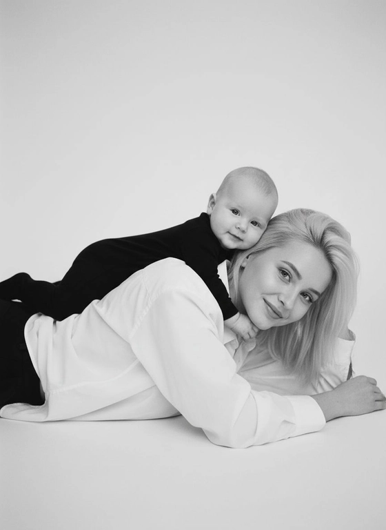 Молодая женщина лежит на полу, на животе, руки вытянуты вверх и окдат на полу, женщина смотрит в камеру, волосы немного закрывают лицо, лицо с мягкой счастливой улыбкой, на её спине лежит маленький  4 месячный ребёнок, обнимая её сверху за шею, он тоже смотрит в камеру и легко улыбается. Женщина одета в белую оверсайз рубашку и черные брюки, рубашка расстегнута у шеи и лежит свободно, ребёнок одет в черный слип. Чёрно‑белая фотография, вертикальный кадр, съёмка в студии на однородном светлом фоне без лишних предметов. Мягкий рассеянный свет, без жёстких теней, ощущение воздушности и легкости. Снято будто на полароид. Без голубых и зеленых оттенков. Атмосфера тёплая, нежная и семейная, передаёт близость и доверие между мамой и ребёнком. Высокое качество, реалистичная детализация кожи, волос и ткани одежды, умеренная глубина резкости, лёгкое виньетирование по краям, акцент на лицах и эмоциях. Стиль – минималистичный семейный портрет, профессиональная студийная съёмка, естественная поза и неподдельные эмоции.

Персонажи занимают 1/4 нижнюю часть кадра, крупный план, женщину видно только по грудь без ног, она лежит боком к камере, камера на расстоянии метра от персонажей. 
Не изменять черты лица и геометрию
