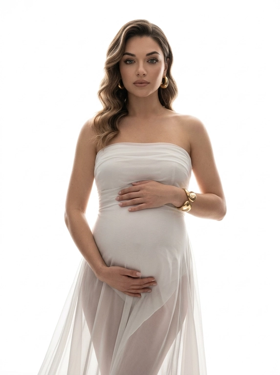 Ultra-realistic high-fashion maternity portrait, framed from knees up. Woman stands centered against a pure white seamless studio background. She wears a strapless sculptural white gown made of sheer layered chiffon and silk, tightly fitted over the pregnant belly and hips, flowing softly downward with translucent folds. The dress emphasizes the round belly clearly and elegantly.

Pose: body slightly angled, both hands gently resting on the belly, one above and one below. Gaze directed straight into the camera, calm, powerful, intimate. Posture upright and confident.

Jewelry minimal: bold gold earrings and a sculptural gold bracelet.

Lighting: high-key studio lighting from behind and slightly from the sides, creating a glowing halo effect around the silhouette, soft rim light outlining the body, gentle frontal fill to keep facial features clear. No harsh shadows, smooth tonal transitions, depth and volume preserved.

Skin looks real and natural with visible texture, no smoothing, no CGI. Editorial luxury maternity aesthetic, timeless, emotional, refined. No text, no logos, no watermark.

Причёска: Hair styled in large polished Hollywood waves with soft volume, falling elegantly over one shoulder.