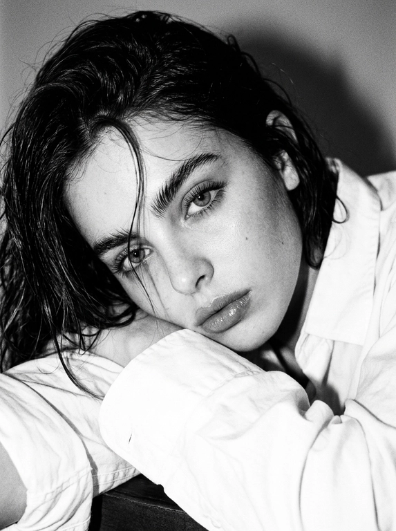 Не меняйте черты лица. Ультрареалистичный черно-белый портрет крупным планом, вертикальный 9:16, снятый с сильной прямой вспышкой на камере, создающий резкие блики и глубокие тени, ярко выраженная эстетика Y2K с более глубоким зернистостью аналоговой пленки и текстурированной монохромной цветоградаданием. Поза идентична: женщина лежит на боку, опираясь головой на сложенные руки на гладкой поверхности, тело слегка наклонено к камере, плотный кадр фокусируется на лице и плечах. Волосы, уложенные в слегка влажную редакционную текстуру с распущенными прядями, падающими по лицу точно так же, как эталон, создавая интимный естественный вид. Макияж точно в соответствии с эталоном: четкие натуральные брови, мягко затененные веки с тонкой подводкой для глаз и длинными натуральными ресницами, гладкая скульптурная кожа с нежным блеском на скулах и переносице, мягко очерченные щеки, натуральные атласные губы с нежным блеском. Идентичный гардероб: белая рубашка оверсайз с расслабленным воротником и видимыми складками ткани. Освещение: сильная вспышка, создающая четкие глянцевые блики на коже и прядях волос, глубокие тени и размерный контраст, высокая детализация кожи и аутентичной текстурой, ярко выраженное наложение зерна пленки Y2K, редакционная фотография красоты.