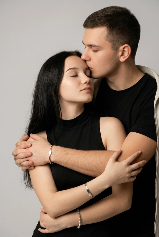 Мужчина и женщина позируют в студии в объятиях. Мужчина стоит позади женщины, обнимая её обеими руками; его тело повернуто к ней, голова слегка наклонена вниз, губы касаются её лба. Женщина немного отклоняет голову назад к его плечу, её глаза закрыты. Кадр — средний, от талии до головы, снят на уровне глаз. Композиция плотная, акцентирует линии рук и соприкосновение лиц. Женщина стоит, прямо обращённая к камере.
На ней облегающий чёрный топ без рукавов с высоким воротом, из плотной матовой ткани.
Мужчина одет в чёрную футболку и чёрную неброскую куртку. На обоих запястьях — тонкие металлические браслеты с минималистичными круглыми подвесками, а женщина носит небольшие светлые металлические серьги с гладкой поверхностью.
Обстановка минималистичная: однотонный светло-серый фон без текстуры.
Освещение — мягкий студийный свет: рассеянный фронтальный и боковой свет создаёт плавные переходы света и тени на лицах и руках. Тени мягкие, средней глубины. Контраст умеренный, без жёстких бликов. Свет равномерно распределён по фигурам, фон подсвечен мягко, без выраженных градиентов.
Съёмка на портретный объектив 85 мм, малая глубина резкости с чётким фокусом на лицах и руках; фон слегка размыт, текстуры кожи и ткани высоко детализированы. Это минималистичный студийный портрет, высокодетализированный и фотореалистичный.
Минималистичное романтическое студийное настроение, нейтральные тёплые тона кожи, низкая–средняя насыщенность, мягкое рассеянное освещение, средний контраст с нежными тенями, плавные переходы бликов, естественная передача кожи, чистый светлый фон, тонкий тональный баланс, современная мягкая редакционная цветокоррекция.

Не изменять мою прическу