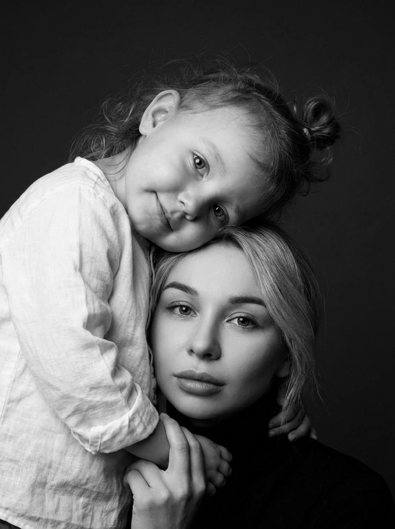 Родитель и ребёнок с внешностью как на приложенных фото, художественная семейная студийная фотосессия. Композиция — вертикальный портрет, крупный план лиц. Родитель внизу смотрит прямо в камеру, лицо фронтально. Ребёнок сверху, голова повернута набок, щека лежит на голове родителя, смотрит в камеру с нежной улыбкой. Рука ребёнка обнимает родителя за шею, видна в кадре, родитель держит руку ребёнка своей рукой.
Одежда: ребёнок в белой льняной рубашке, родитель в чёрной водолазке. 
Фон — тёмно-серый почти чёрный однотонный студийный, создаёт контраст. Освещение — мягкое боковое студийное, создаёт объём и глубину.
Чёрно-белая художественная фотография с выразительными контрастами. Атмосфера нежная, интимная, спокойная.